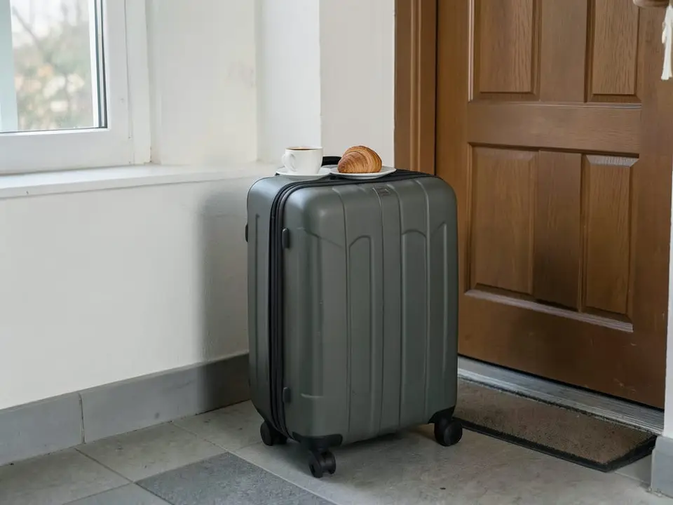 A packed carry-on suitcase stands by an apartment door next to a small pastry and coffee cup, reflecting little treats and last-minute trips. AI-generated editorial illustration; not a documentary photograph