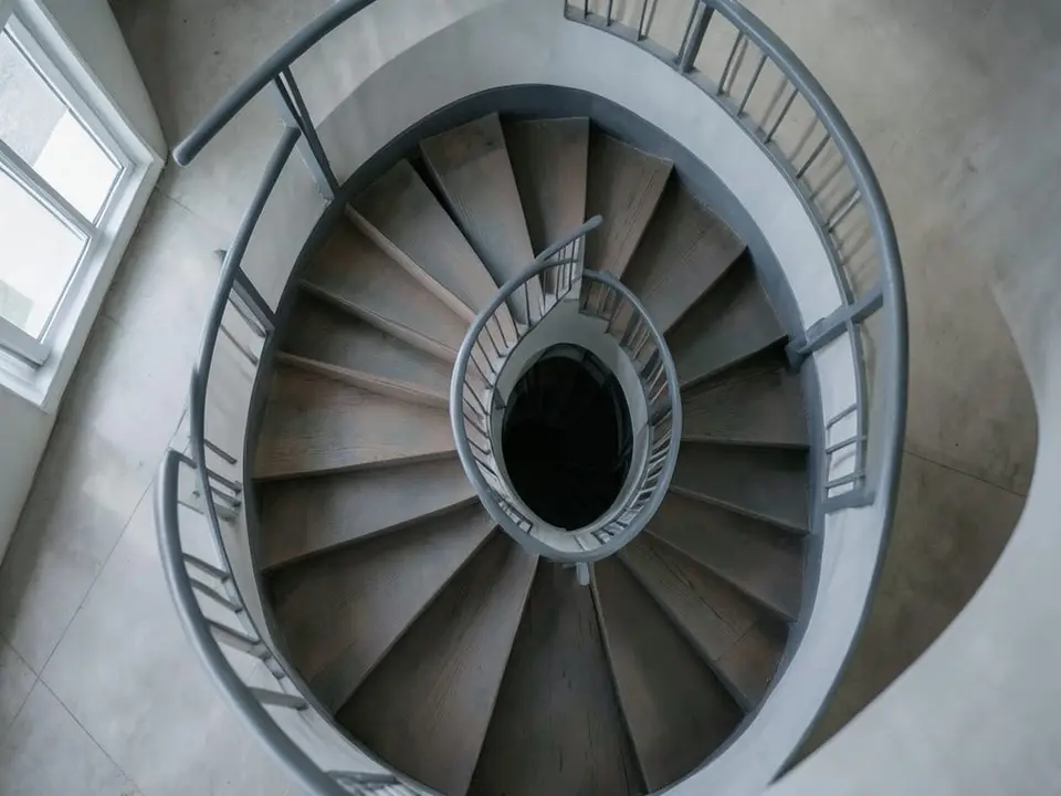 A spiral staircase winding tightly downward from above, representing a debt spiral in which interest rates on new Treasury bonds and notes at around 5% outpace medium-term nominal economic growth expected to be closer to 4%. AI-generated editorial illustration; not a documentary photograph
