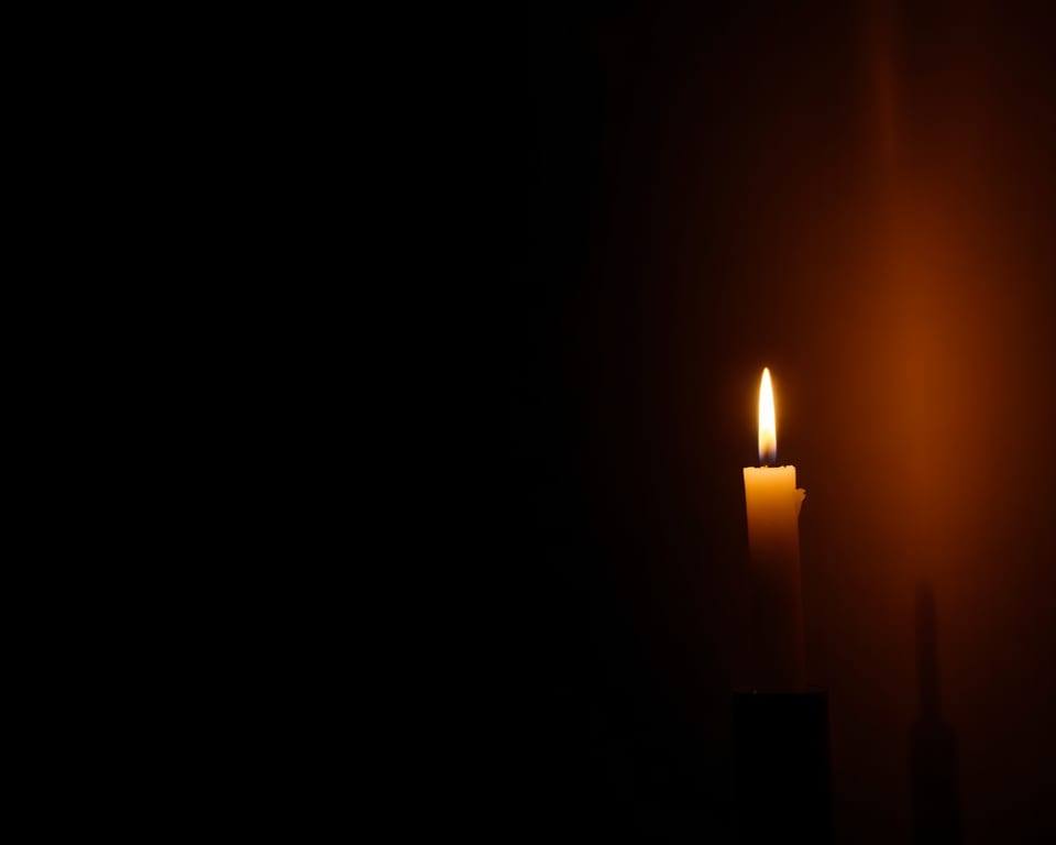 a single lit candle glowing warmly in a dark quiet room AI-generated editorial illustration; not a documentary photograph
